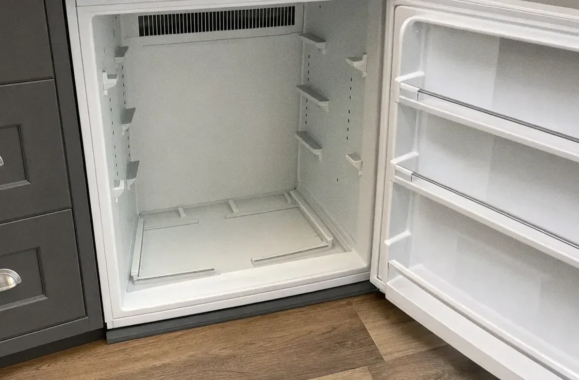White refrigerator cabinet standing open on empty shelves and door bins above a wood floor
